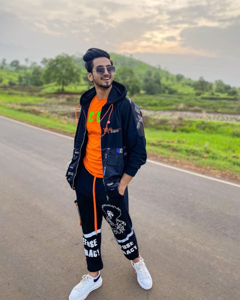 Faisal Shaikh posing in front of camera wearing black jacket  and funky trouscer, orange tshirt and shades
