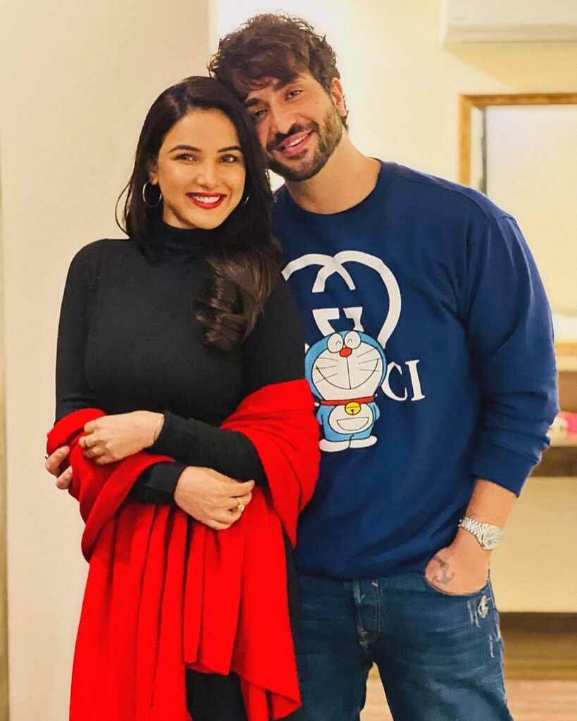 Smiling Aly Goni in blue t-shirt and Jasmin Bhasin in black high neck top and red stole posing for camera - live in relationship India 