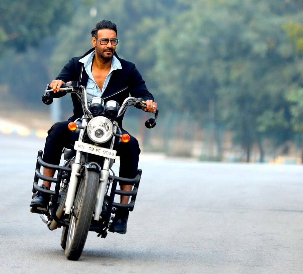Ajay Devgan in black suit and light color shirt riding an bike and posing for camera and showing off his Pompadour hairstyle - face shape