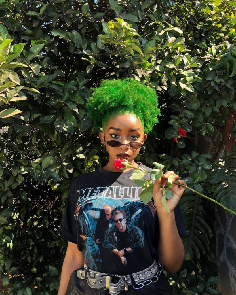 Girl in black printed t-shirt with goggles holding a rose and showing her leafy greens hair color - green hair color for women