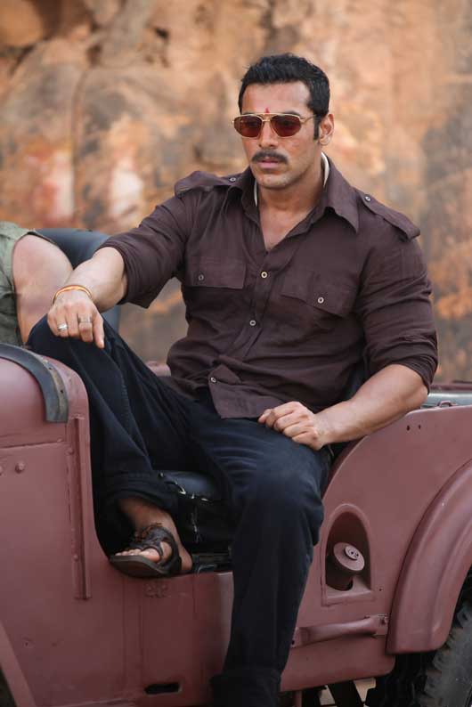 John Abraham in black trouser with brown shirt and goggles sitting on a car seat and posing for camera - Indian actors Latest Hairstyles