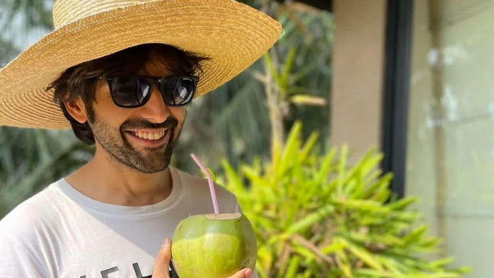 Smiling Kartik Aaryan in white round neck t-shirt with a jute hat and googles drinking coconut water - Kartik Aaryan hair colour